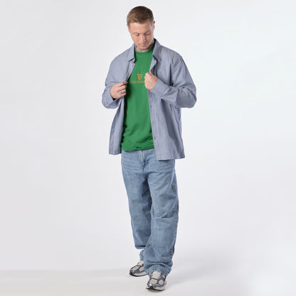 A man stands against a plain white background, wearing the Guinness Gilroy Toucan Tee in green from Guinness Webstore US, an unbuttoned blue shirt, loose blue jeans, and gray sneakers, looking down while adjusting his shirt.