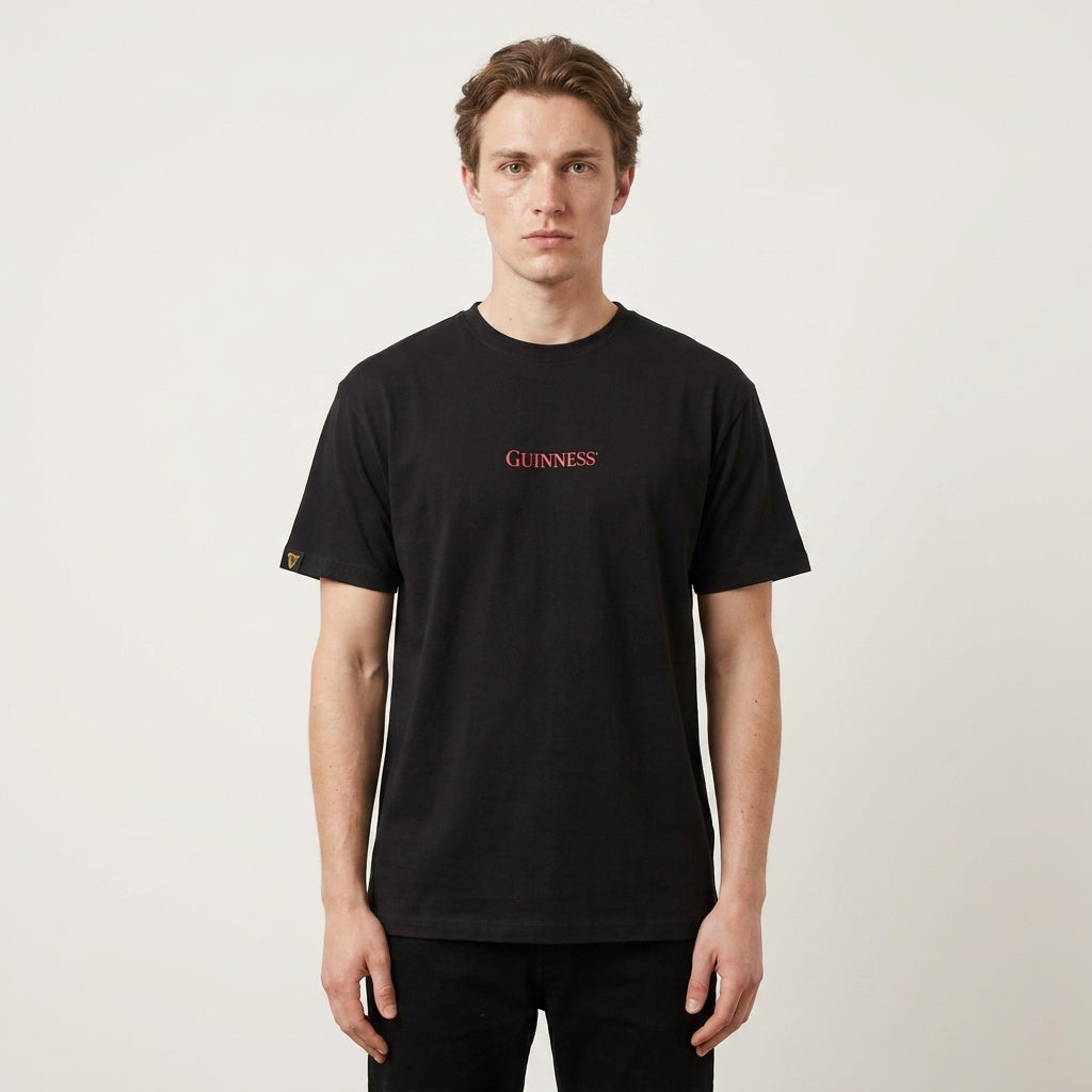 A person stands facing forward, wearing a Guinness Gilroy Ostrich Tee – Black from Guinness Webstore US, featuring "Guinness" in red text on the chest, against a plain white background.