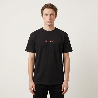 A person stands facing forward, wearing a Guinness Gilroy Ostrich Tee – Black from Guinness Webstore US, featuring "Guinness" in red text on the chest, against a plain white background.