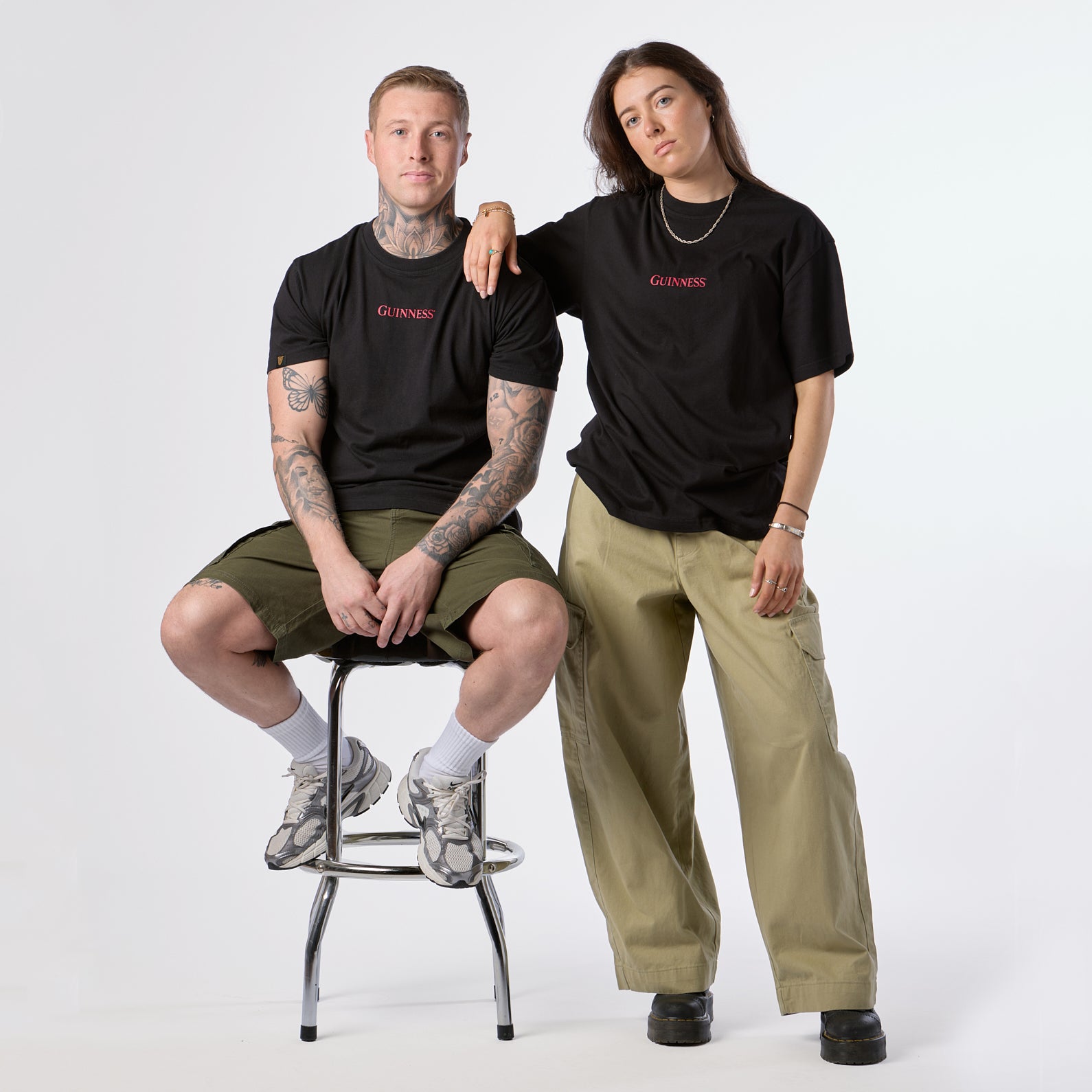 Two people pose in casual outfits from the iconic Gilroy collection; both wear the limited edition Guinness Gilroy Ostrich Tee in black from Guinness Webstore US, paired with olive green pants. One sits on a stool while the other stands with an arm resting on the seated person’s shoulder.