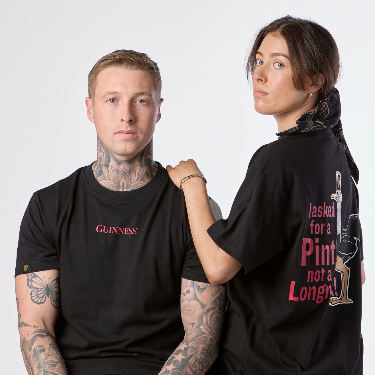 A tattooed man wearing the Guinness Gilroy Ostrich Tee – Black from the Guinness Webstore US, a limited edition piece inspired by the iconic Gilroy collection, sits beside a woman who stands next to him in a black t-shirt emblazoned with the phrase “I asked for a pint, not a long.” Both face the camera.