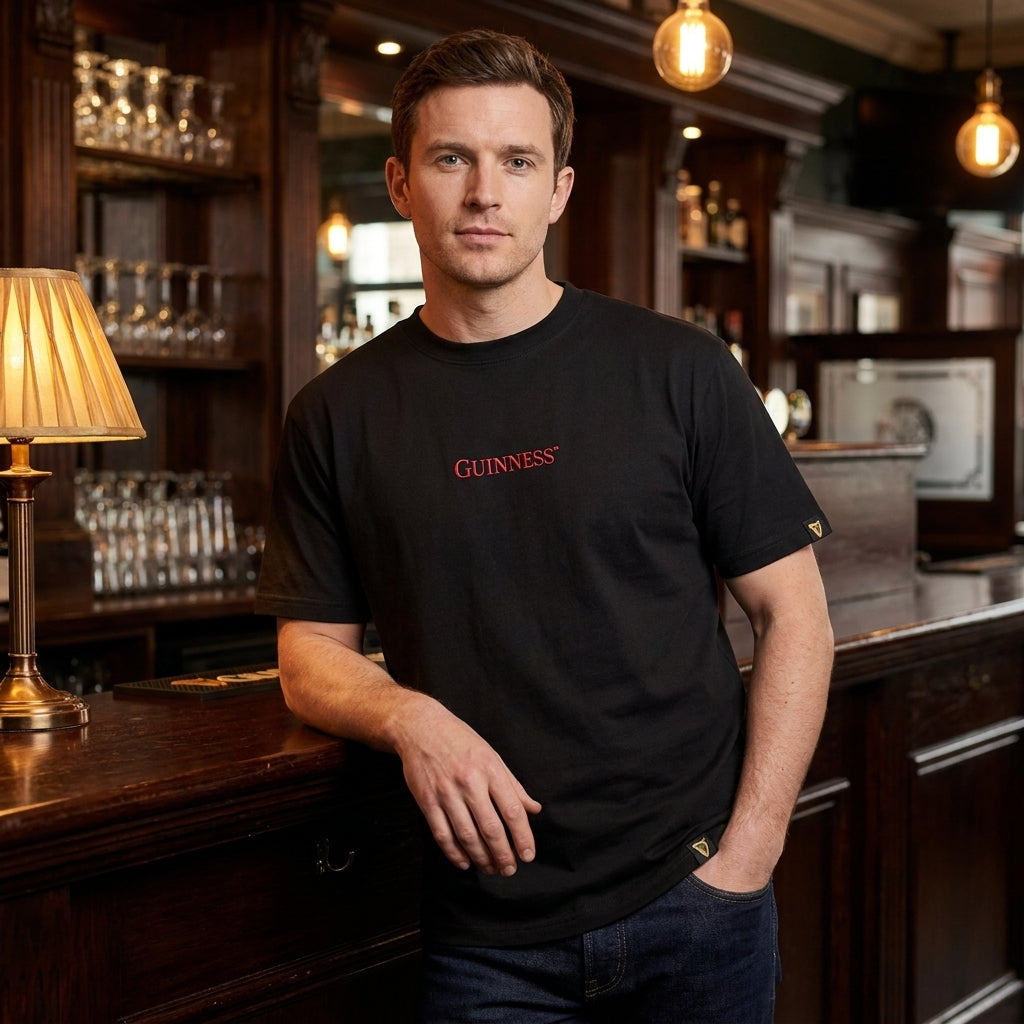 A man in a Guinness Gilroy Ostrich Tee – Black from Guinness Webstore US stands at a wooden bar, with shelves of glasses and warm lighting in the background.