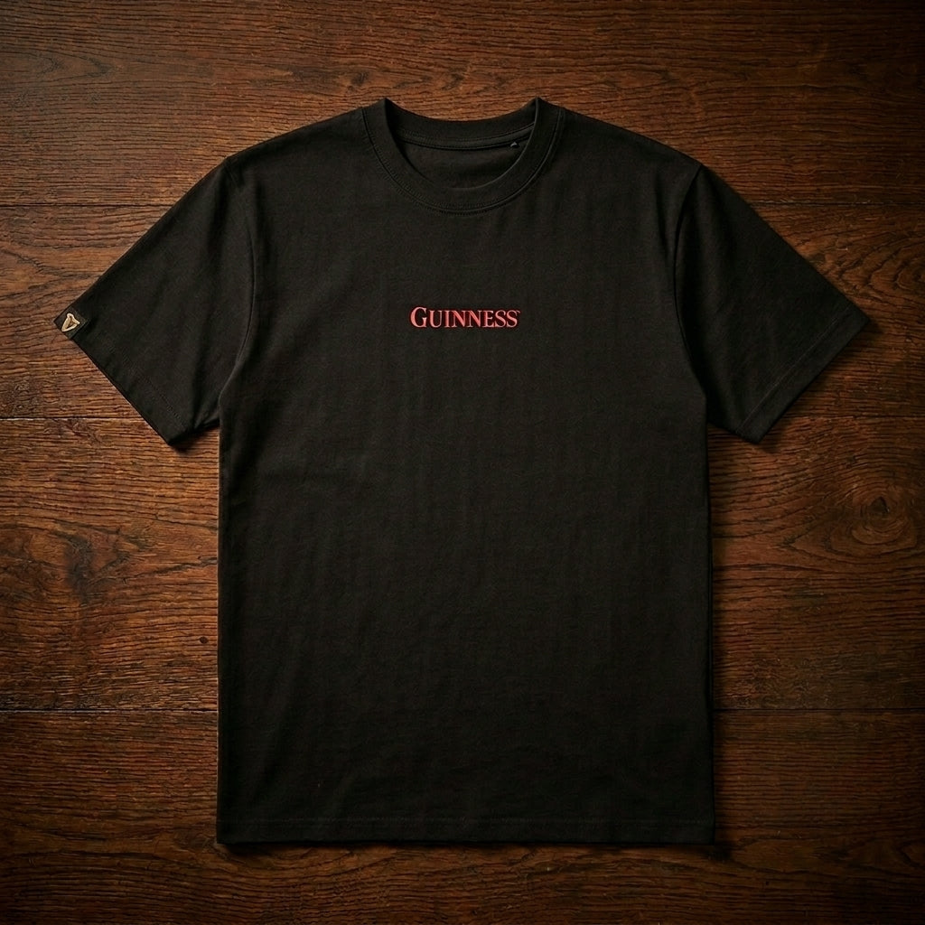 The Guinness Webstore US features the limited edition Guinness Gilroy Ostrich Tee in black, with "GUINNESS" in red on the chest, displayed flat on a dark wooden surface.