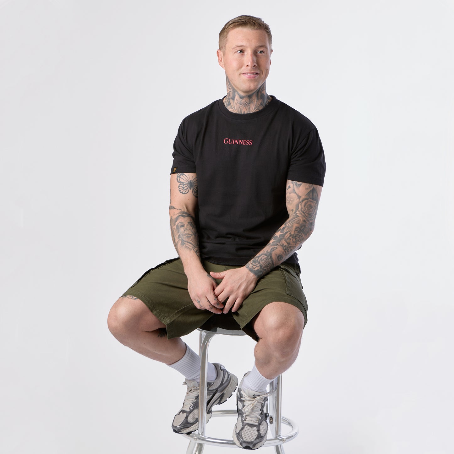 A man with tattoos sits on a metal stool wearing the Guinness Gilroy Ostrich Tee in black from the Guinness Webstore US, paired with olive shorts, white socks, and sneakers against a plain white background.