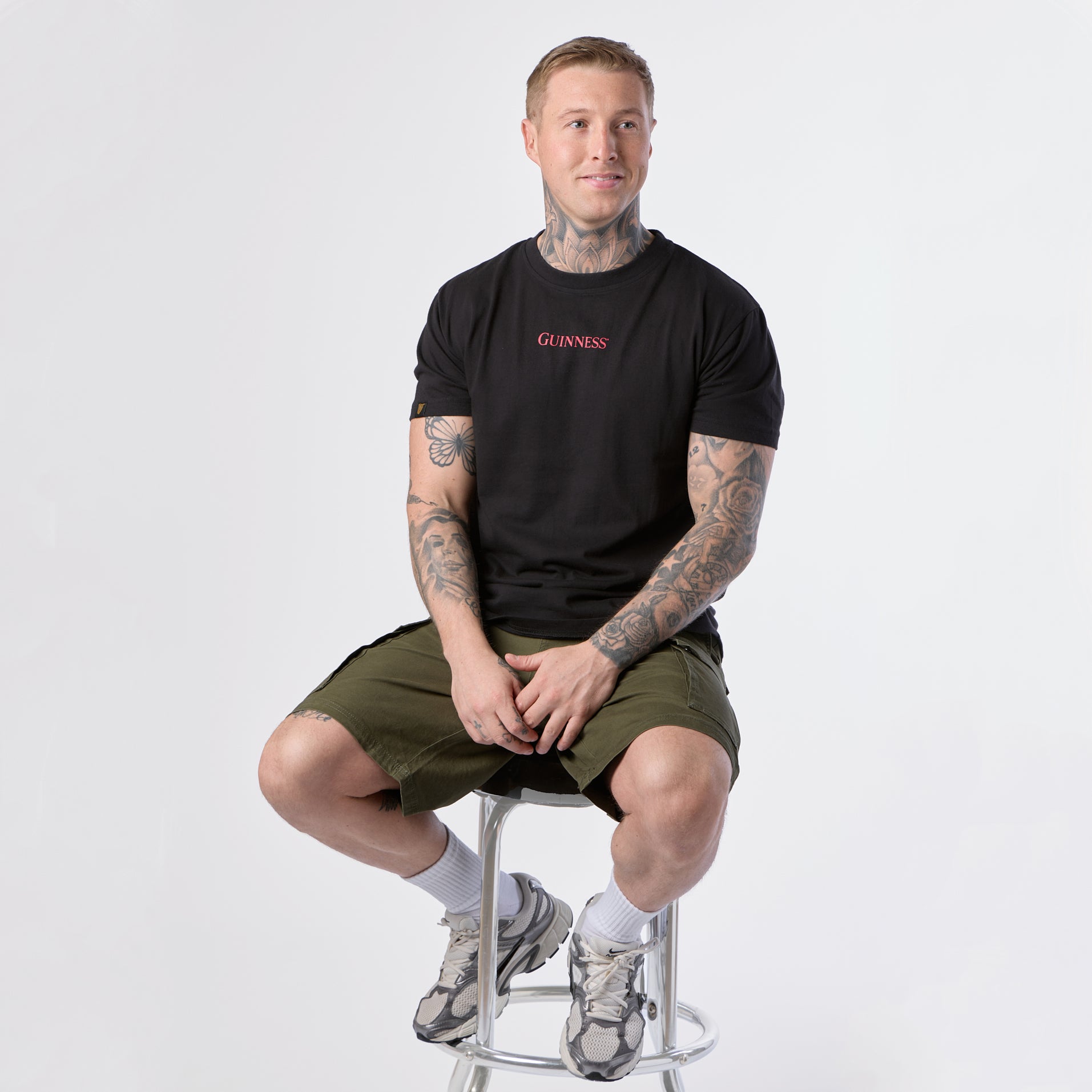 A man with tattoos sits on a metal stool wearing the Guinness Gilroy Ostrich Tee in black from the Guinness Webstore US, paired with olive shorts, white socks, and sneakers against a plain white background.