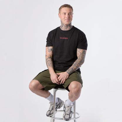 A man with tattoos sits on a metal stool wearing the Guinness Gilroy Ostrich Tee in black from the Guinness Webstore US, paired with olive shorts, white socks, and sneakers against a plain white background.