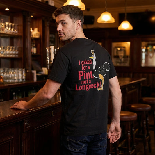 A man stands at a wooden bar wearing the Guinness Gilroy Ostrich Tee – Black from Guinness Webstore US, featuring “I asked for a Pint, not a Longneck!” printed in red and white on the back.