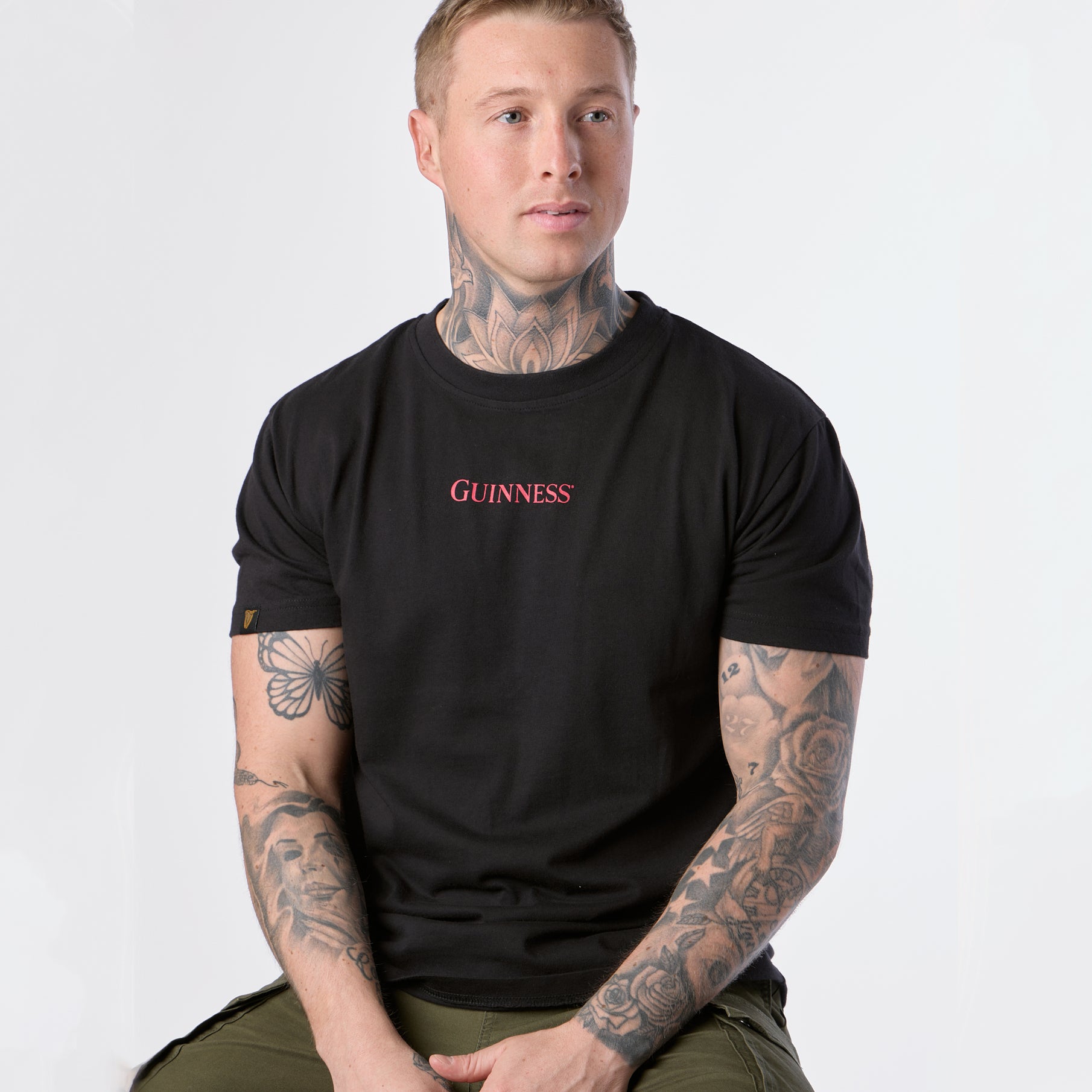 A tattooed man with short hair sits slightly turned to the side, wearing the black Guinness Gilroy Ostrich Tee from Guinness Webstore US and green pants, set against a plain white background.