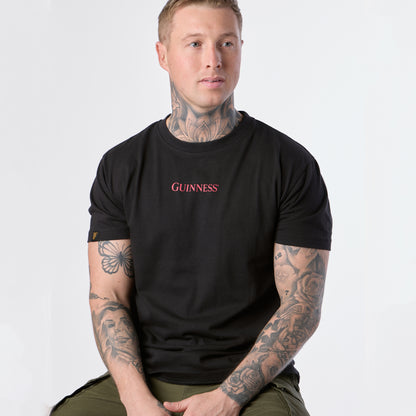 A tattooed man with short hair sits slightly turned to the side, wearing the black Guinness Gilroy Ostrich Tee from Guinness Webstore US and green pants, set against a plain white background.