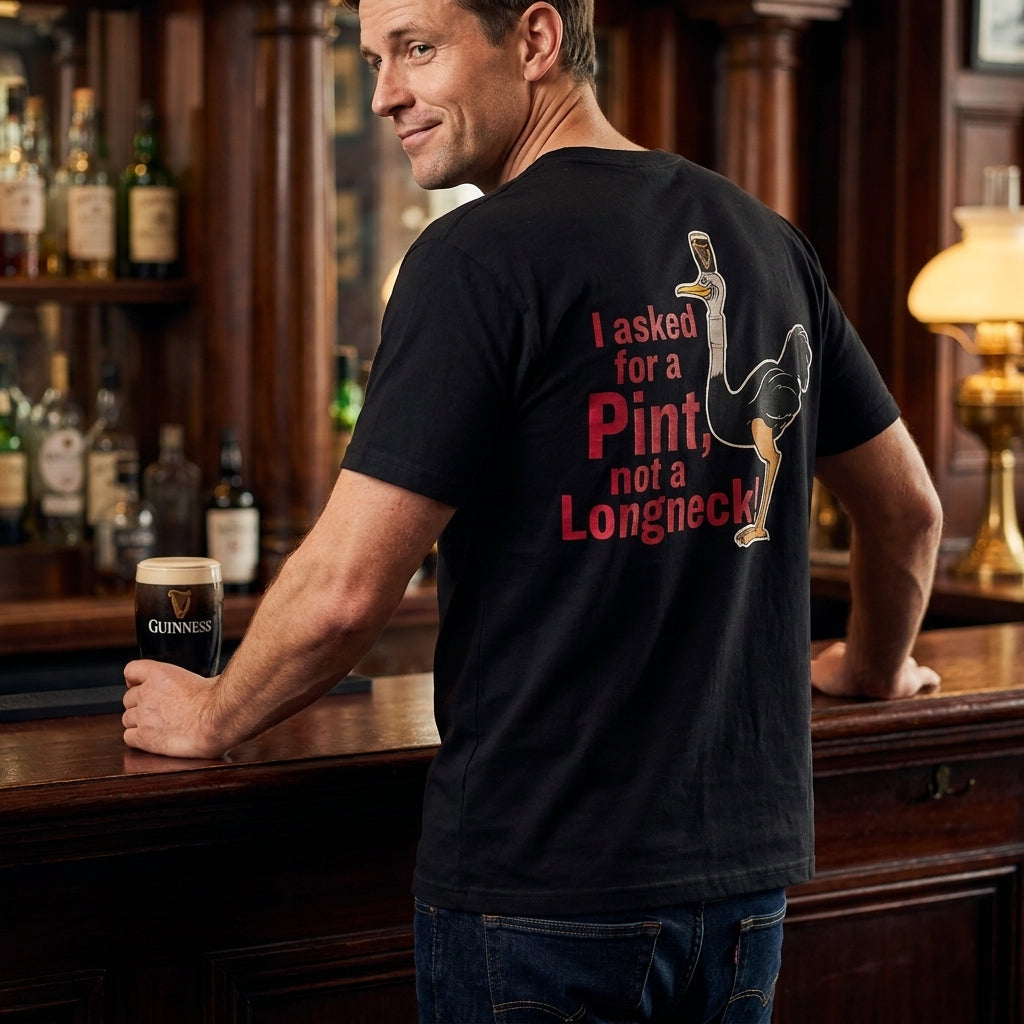 At a bar, a man holds a pint of Guinness and wears the Guinness Gilroy Ostrich Tee – Black from Guinness Webstore US, featuring “I asked for a Pint, not a Longneck!” and an ostrich with a pint glass from the iconic Gilroy collection.