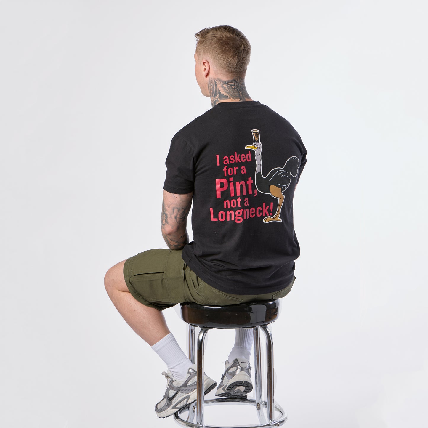 A man sits on a stool facing away, wearing the black Guinness Gilroy Ostrich Tee from Guinness Webstore US, featuring the limited edition Iconic Gilroy collection design with the text “I asked for a Pint, not a Longneck!”, paired with khaki shorts against a white background.