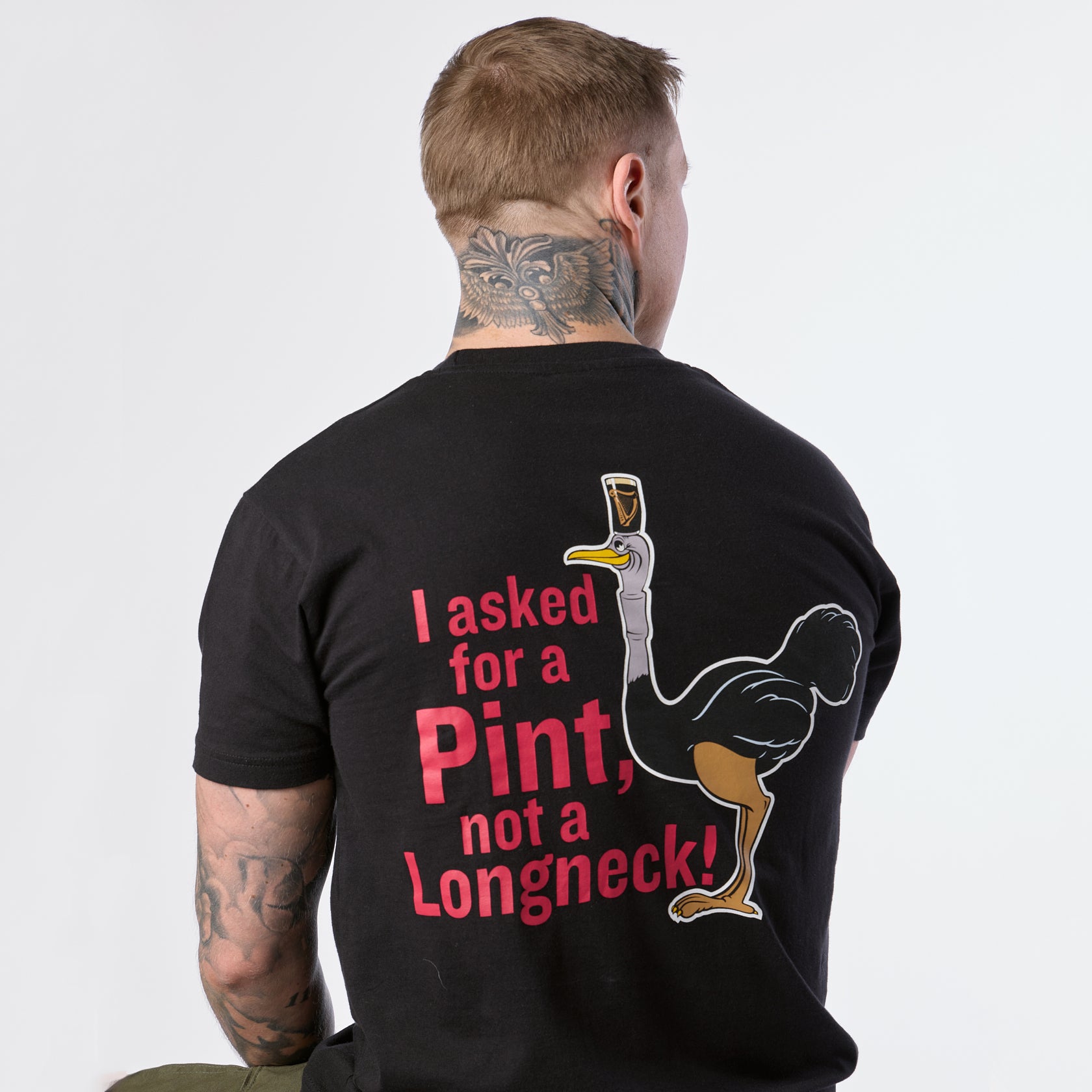 A person with neck tattoos is wearing the limited edition Guinness Gilroy Ostrich Tee – Black from Guinness Webstore US, featuring an ostrich graphic and the phrase, “I asked for a Pint, not a Longneck!”.