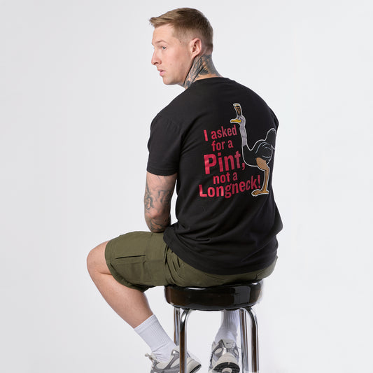 A man sits on a stool, turned to display the back of his black Guinness Gilroy Ostrich Tee from Guinness Webstore US, featuring the phrase “I asked for a Pint, not a Longneck!” alongside an illustration of a beer glass—part of the iconic Gilroy collection.