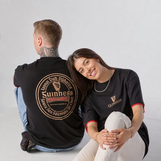 A man with tattoos sits with his back to the camera while a woman beside him smiles at the camera, both wearing the Guinness Irish Label Black Tee from Guinness Webstore US against a plain white background.