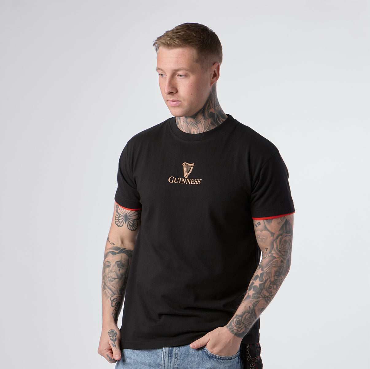 A person with tattoos wears a Guinness Irish Label Black Tee from Guinness Webstore US, featuring red trim on the sleeves, paired with blue jeans and standing against a plain light background.