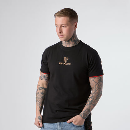 A person with tattoos wears a Guinness Irish Label Black Tee from Guinness Webstore US, featuring red trim on the sleeves, paired with blue jeans and standing against a plain light background.