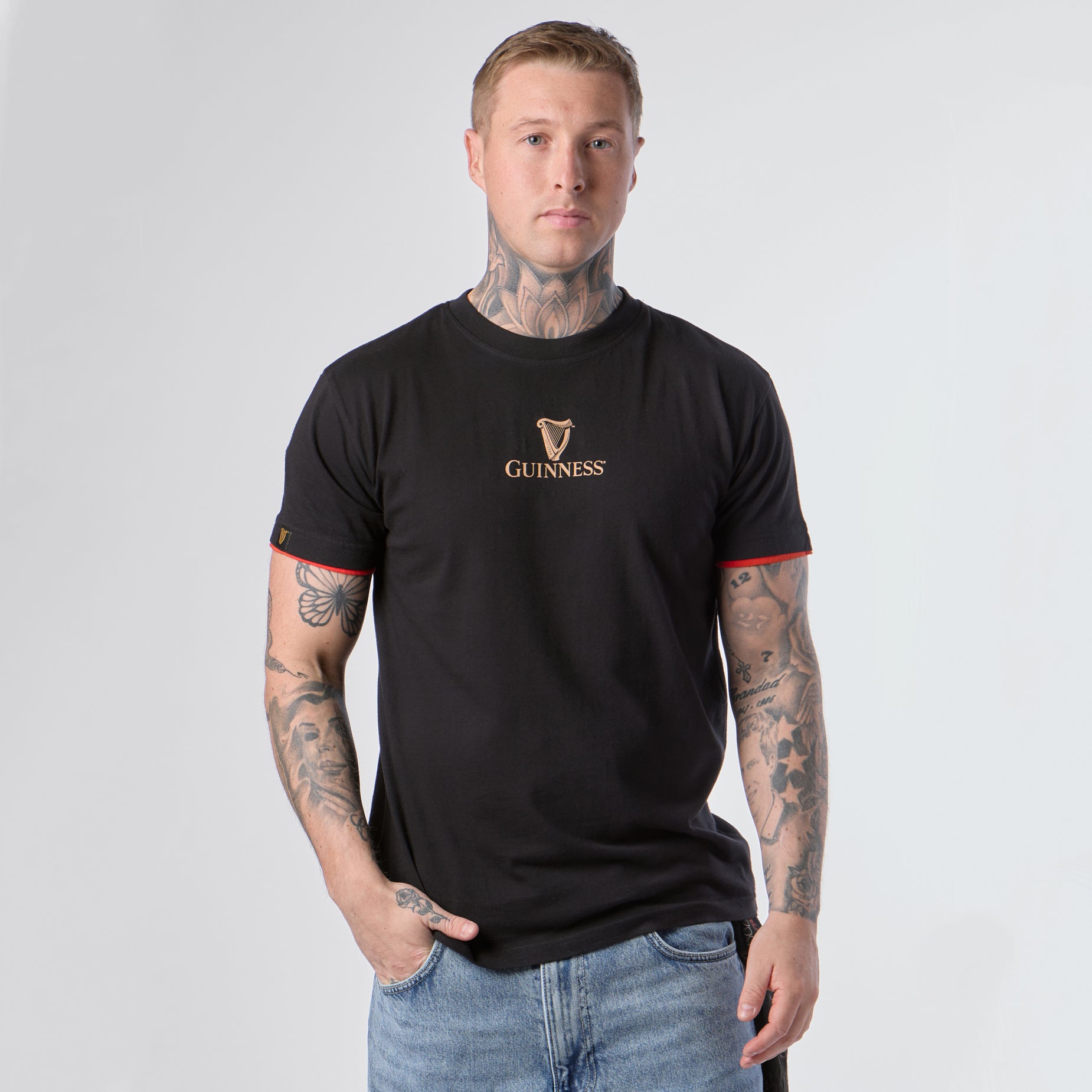 A man with tattoos wears the Guinness Irish Label Black Tee from Guinness Webstore US and blue jeans, standing against a plain light background, facing forward with one hand in his pocket.