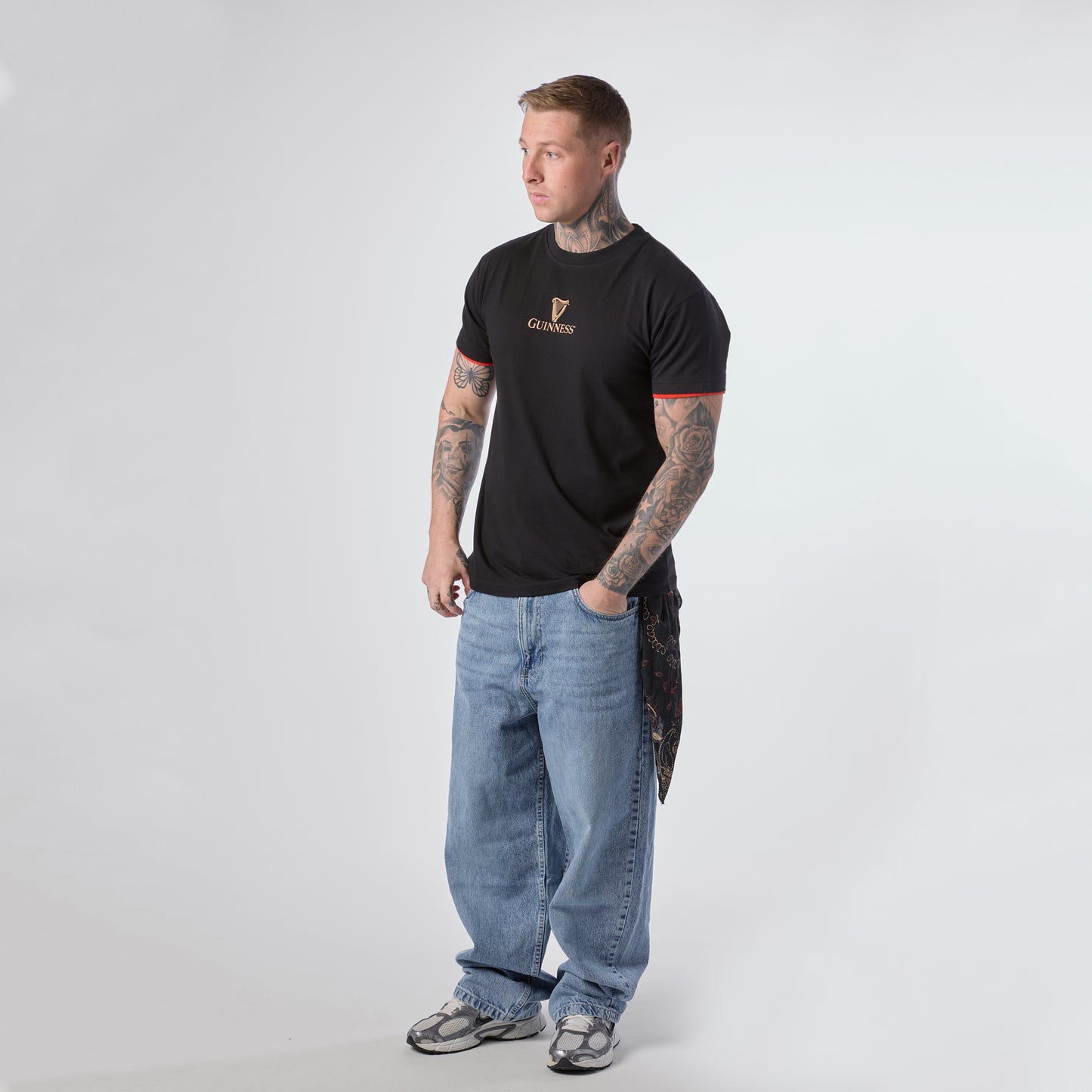 A man with tattoos stands facing slightly left, wearing a Guinness Irish Label Black Tee from Guinness Webstore US, paired with baggy blue jeans, gray sneakers, and a bandana hanging from his pocket against a plain white background.