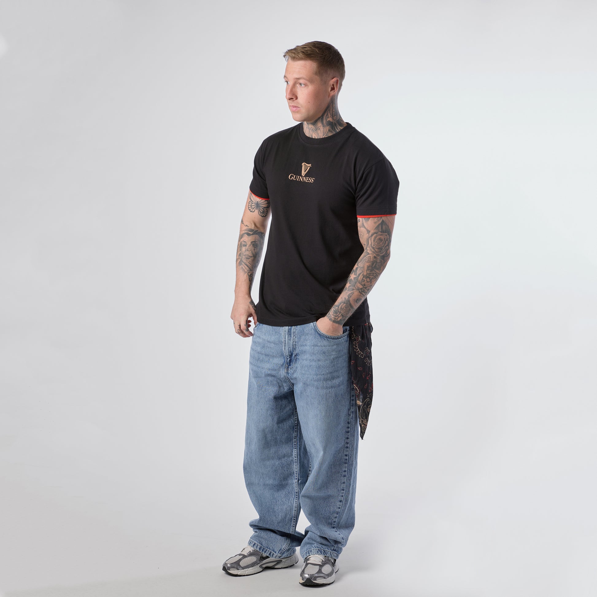 A man with tattoos stands facing slightly left, wearing a Guinness Irish Label Black Tee from Guinness Webstore US, paired with baggy blue jeans, gray sneakers, and a bandana hanging from his pocket against a plain white background.