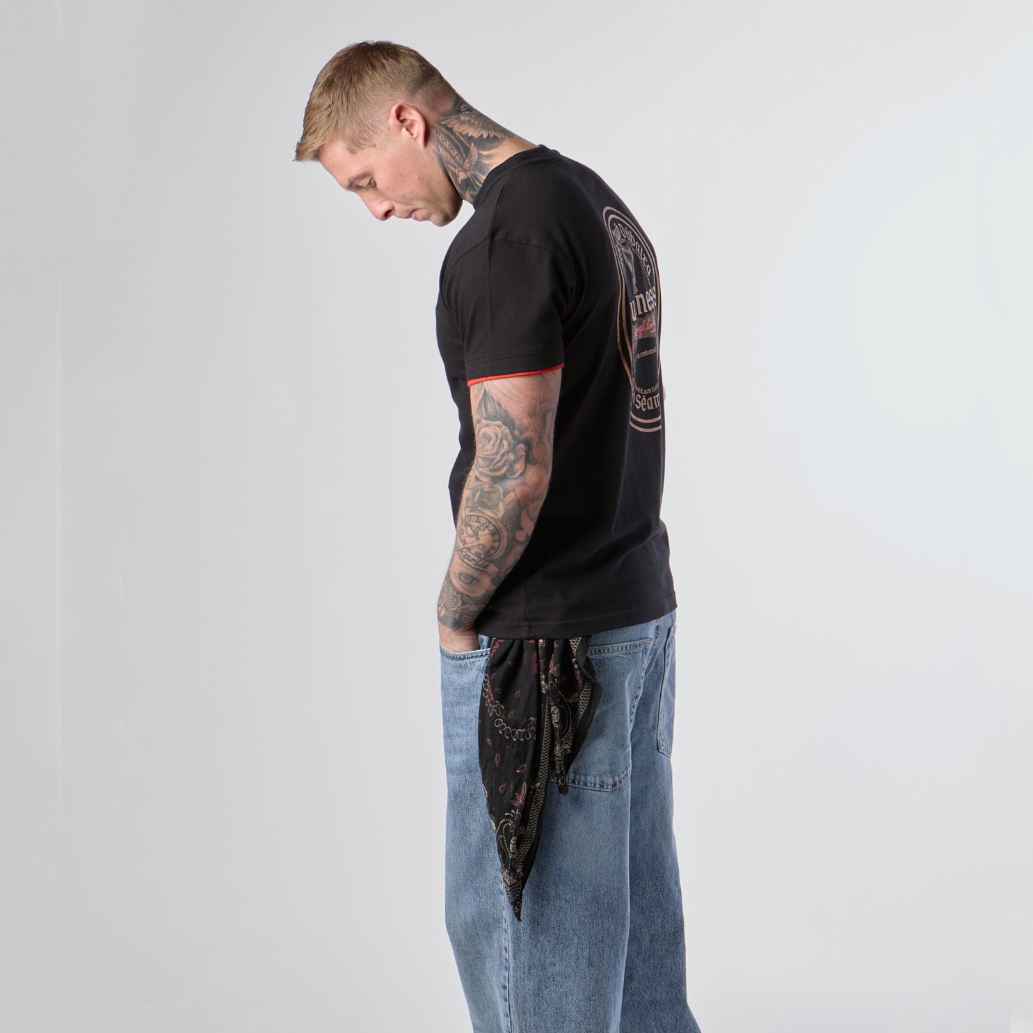 A tattooed man stands in profile, looking down, wearing the Guinness Webstore US Guinness Irish Label Black Tee featuring a logo on the back, paired with blue jeans and a bandana hanging from his pocket against a plain light background.