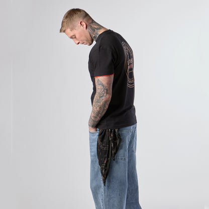 A tattooed man stands in profile, looking down, wearing the Guinness Webstore US Guinness Irish Label Black Tee featuring a logo on the back, paired with blue jeans and a bandana hanging from his pocket against a plain light background.