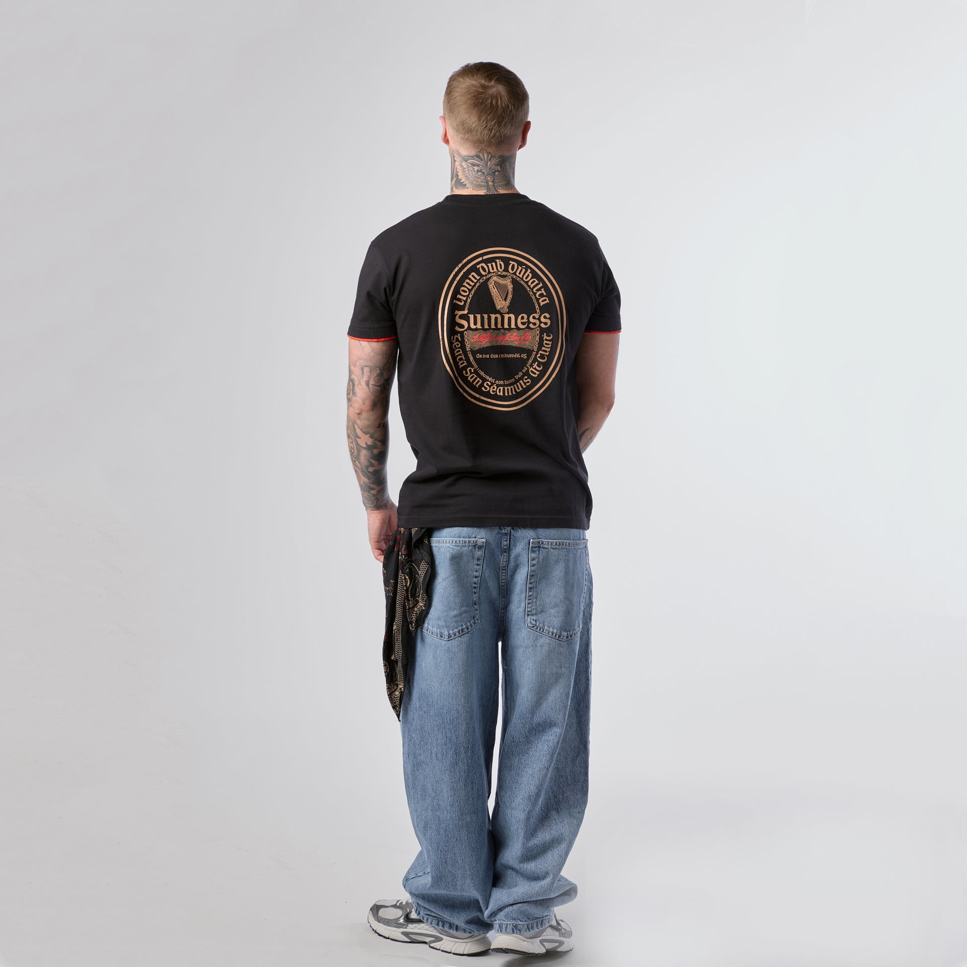 A man with tattoos stands facing away, wearing a Guinness Irish Label Black Tee from the Guinness Webstore US, paired with blue jeans, sneakers, and holding a patterned cloth in his left hand.