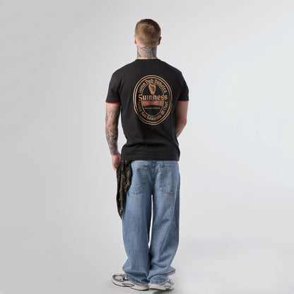 A man with tattoos stands facing away, wearing a Guinness Irish Label Black Tee from the Guinness Webstore US, paired with blue jeans, sneakers, and holding a patterned cloth in his left hand.