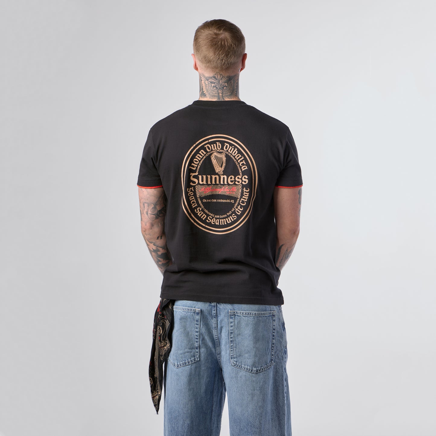A tattooed individual stands facing away, wearing the Guinness Irish Label Black Tee from Guinness Webstore US, which showcases a bold circular Guinness logo design on the back, paired with loose blue jeans.