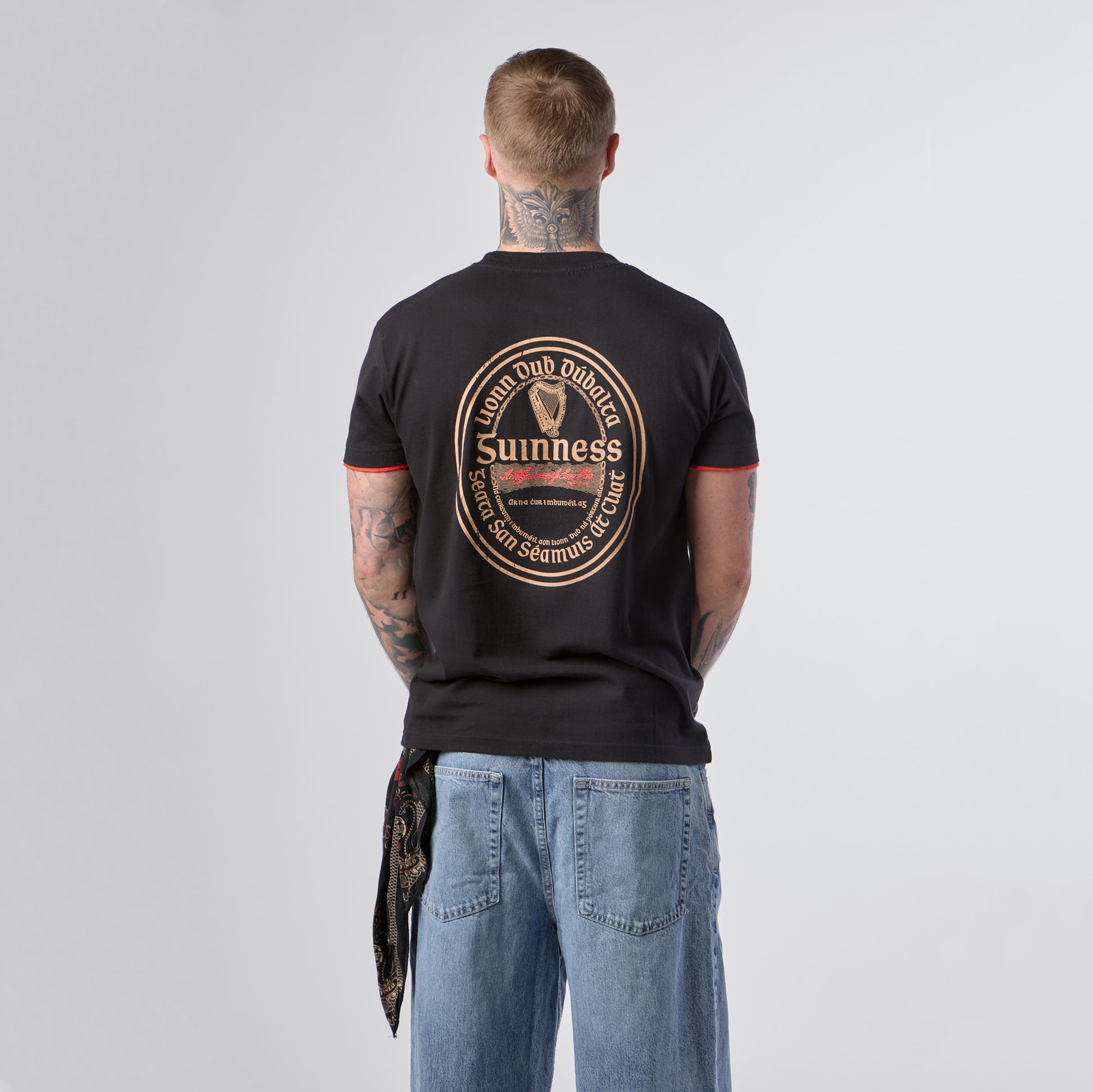 A tattooed individual stands facing away, wearing the Guinness Irish Label Black Tee from Guinness Webstore US, which showcases a bold circular Guinness logo design on the back, paired with loose blue jeans.