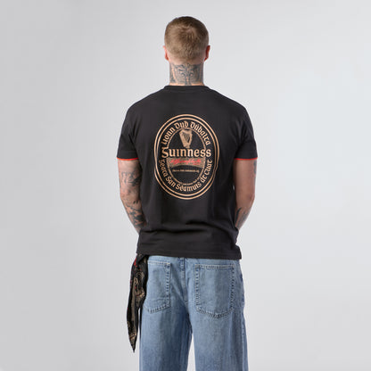 A tattooed individual stands facing away, wearing the Guinness Irish Label Black Tee from Guinness Webstore US, which showcases a bold circular Guinness logo design on the back, paired with loose blue jeans.