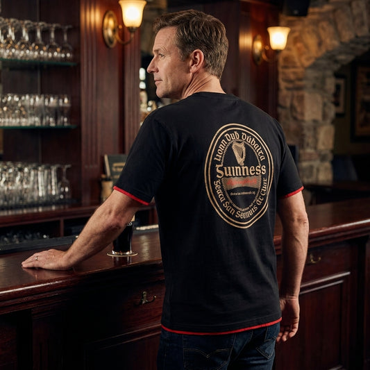 A man at a bar rests his hand on the counter, wearing the Guinness Gaelic Label Black Tee from Guinness Webstore US, which features a circular "Sunness" logo on the back.