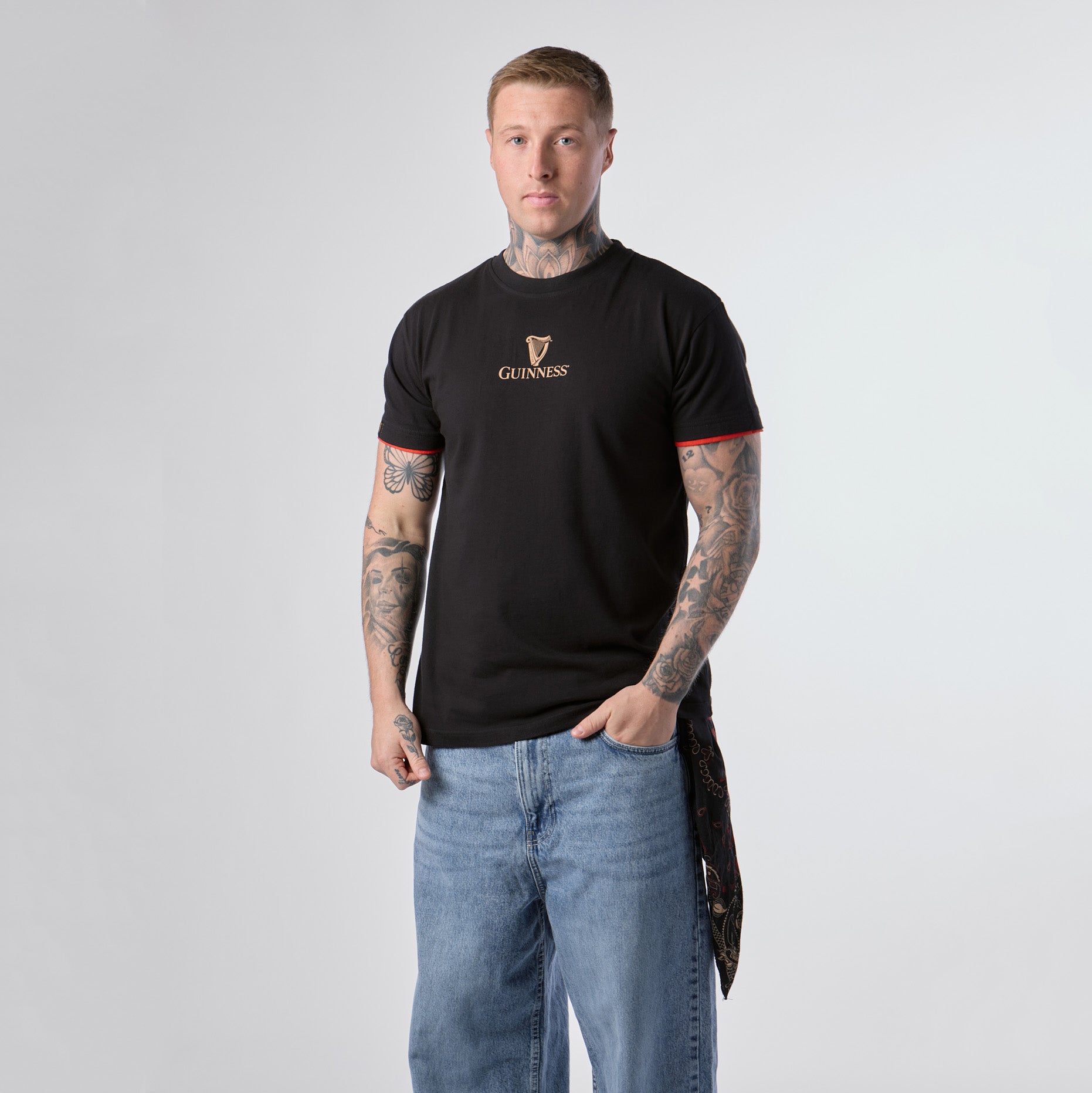 A person with tattoos stands against a plain white background, wearing the Guinness Irish Label Black Tee from Guinness Webstore US and blue jeans, with one hand in their pocket.