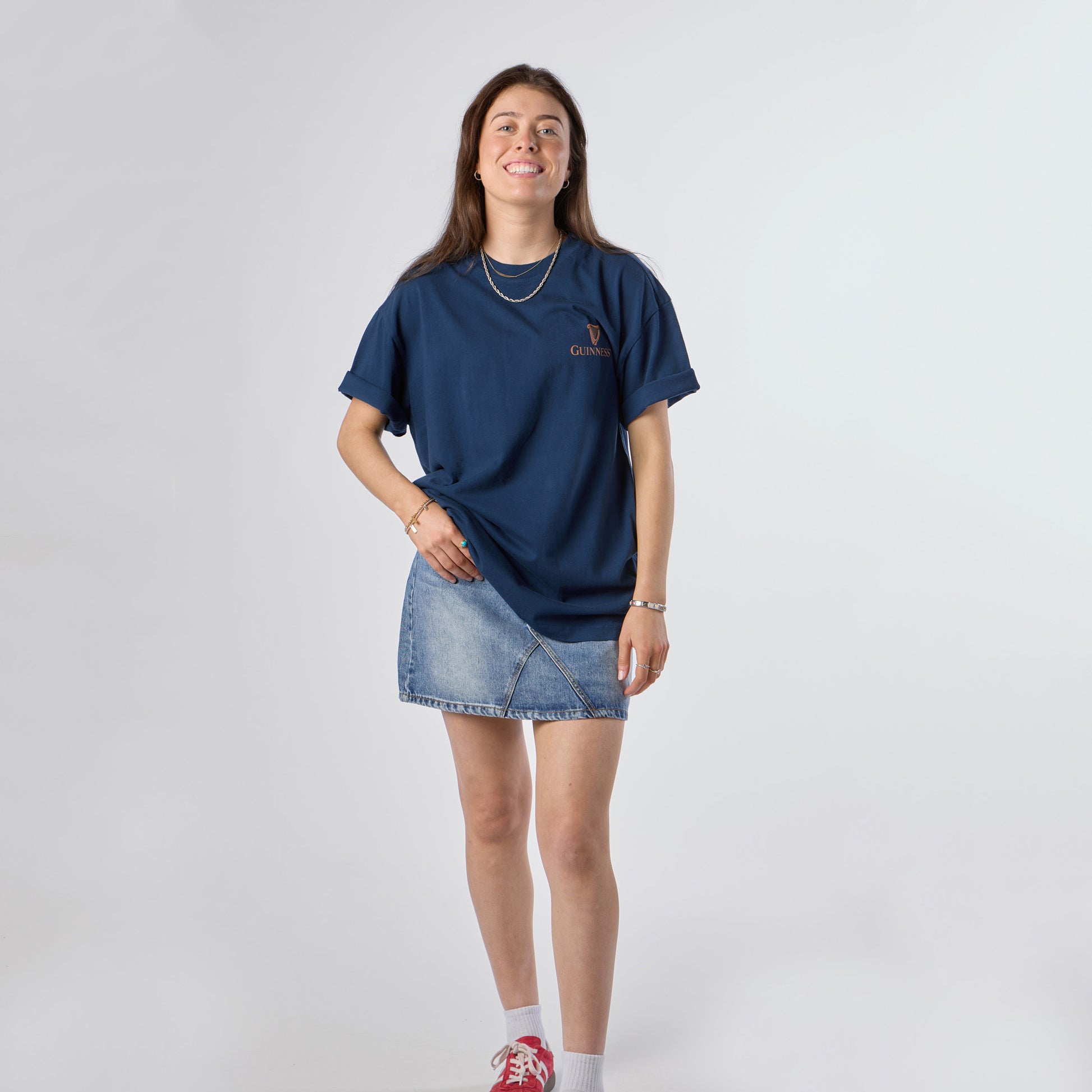 A woman stands and smiles against a plain light background, wearing the Guinness Gilroy Seal Tee – Blue from Guinness Webstore US, paired with a denim skirt, white socks, and red sneakers.