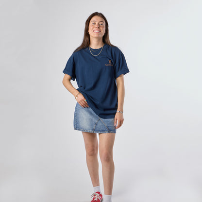 A woman stands and smiles against a plain light background, wearing the Guinness Gilroy Seal Tee – Blue from Guinness Webstore US, paired with a denim skirt, white socks, and red sneakers.