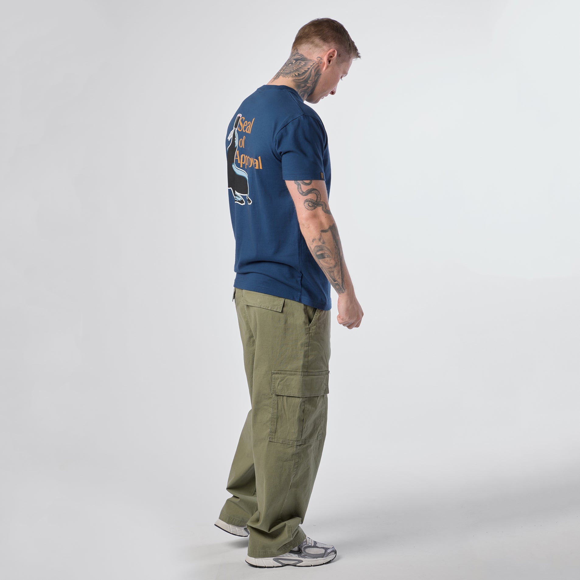 A man with tattoos wears the Guinness Gilroy Seal Tee – Blue from the Guinness Webstore US, paired with olive cargo pants and gray sneakers, standing sideways against a plain white background.