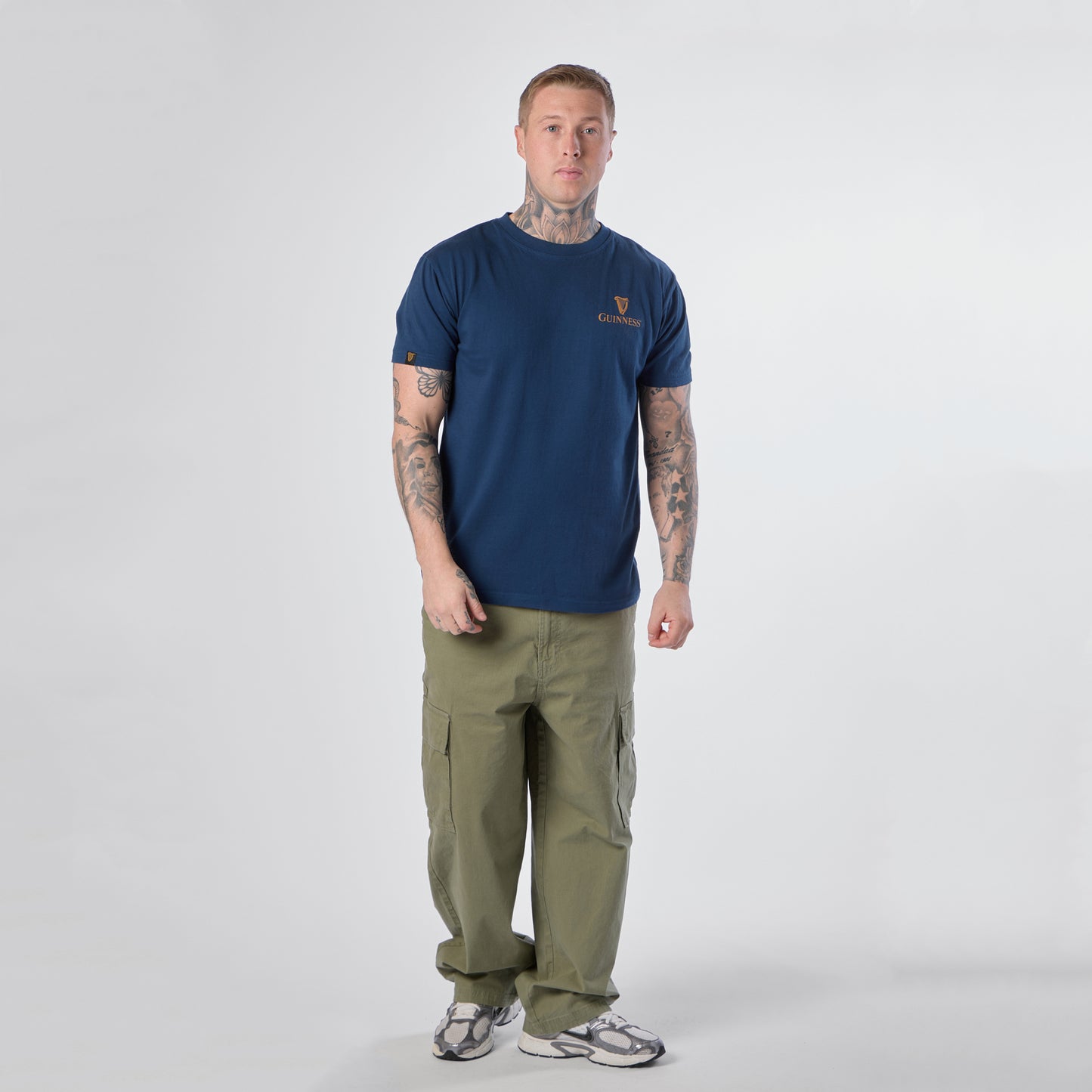 A man with tattoos stands facing forward, wearing a Guinness Gilroy Seal Tee in blue from the Guinness Webstore US, paired with olive cargo pants and white sneakers against a plain white background.