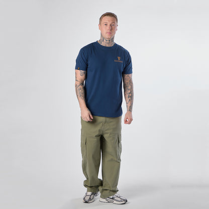 A man with tattoos stands facing forward, wearing a Guinness Gilroy Seal Tee in blue from the Guinness Webstore US, paired with olive cargo pants and white sneakers against a plain white background.