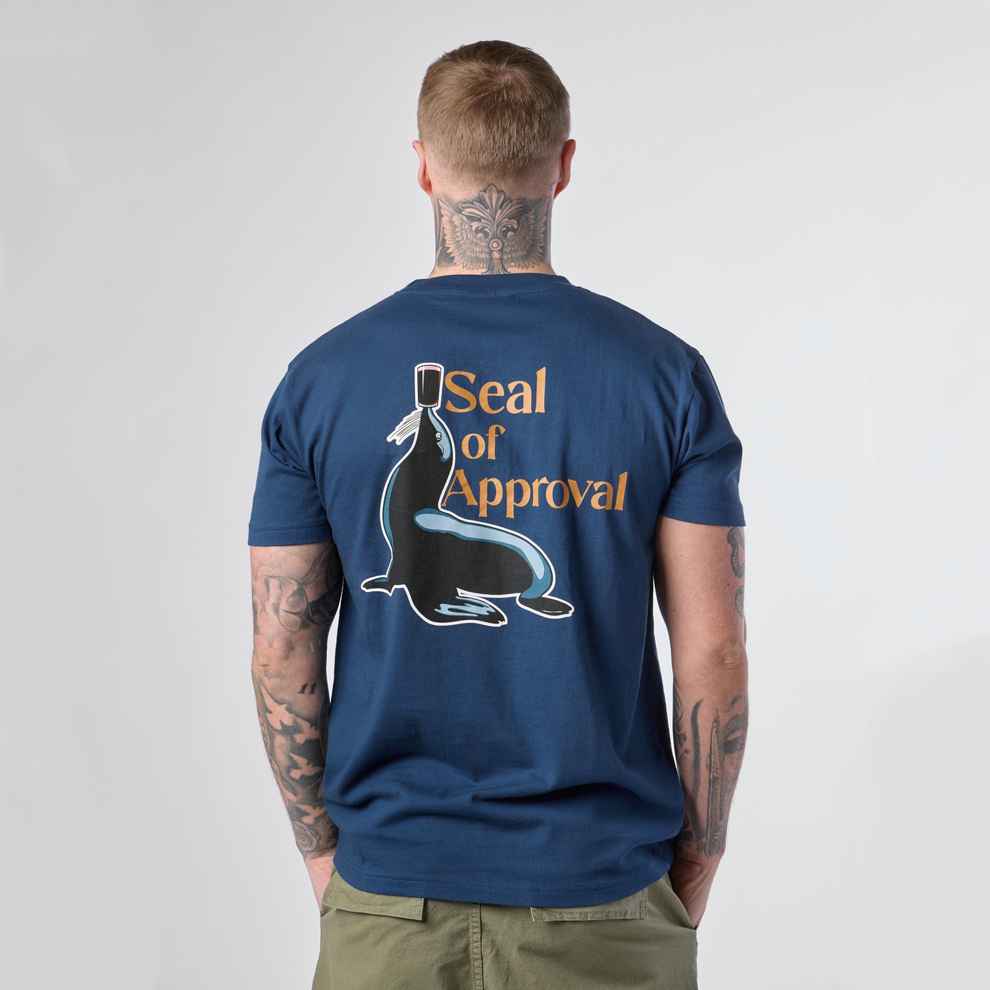 A person seen from behind wears the Guinness Gilroy Seal Tee – Blue from Guinness Webstore US, featuring a graphic of a seal balancing a stamp and the words “Seal of Approval.”.