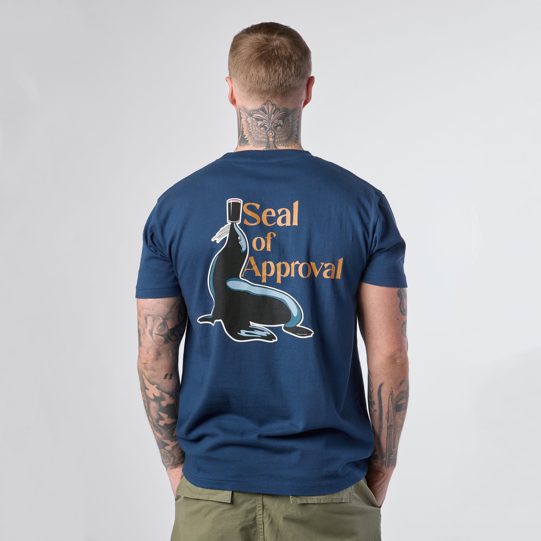 A person seen from behind wears the Guinness Gilroy Seal Tee – Blue from Guinness Webstore US, featuring a graphic of a seal balancing a stamp and the words “Seal of Approval.”.