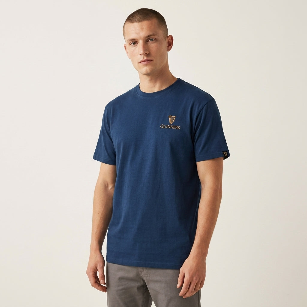 A man in a Guinness Gilroy Seal Tee – Blue from Guinness Webstore US and gray pants stands against a plain light background.