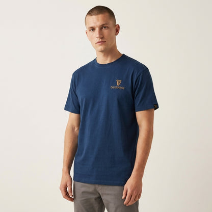 A man in a Guinness Gilroy Seal Tee – Blue from Guinness Webstore US and gray pants stands against a plain light background.