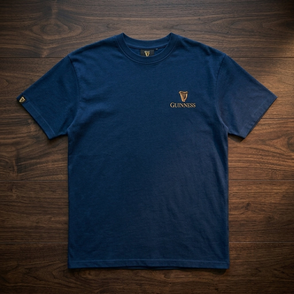 The Guinness Gilroy Seal Tee – Blue by Guinness Webstore US, featuring a gold logo on the left chest, is laid flat on a dark wooden surface.