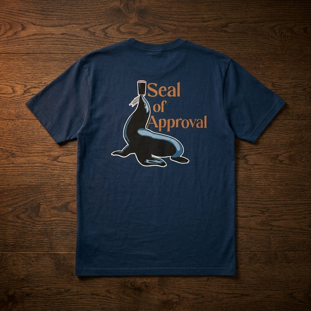 The Guinness Gilroy Seal Tee – Blue from Guinness Webstore US is displayed on a wooden surface, featuring a seal with a pint of Guinness balanced on his nose and the text “Seal of Approval” on the back.