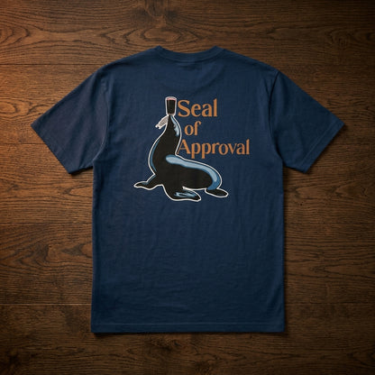 The Guinness Gilroy Seal Tee – Blue from Guinness Webstore US is displayed on a wooden surface, featuring a seal with a pint of Guinness balanced on his nose and the text “Seal of Approval” on the back.