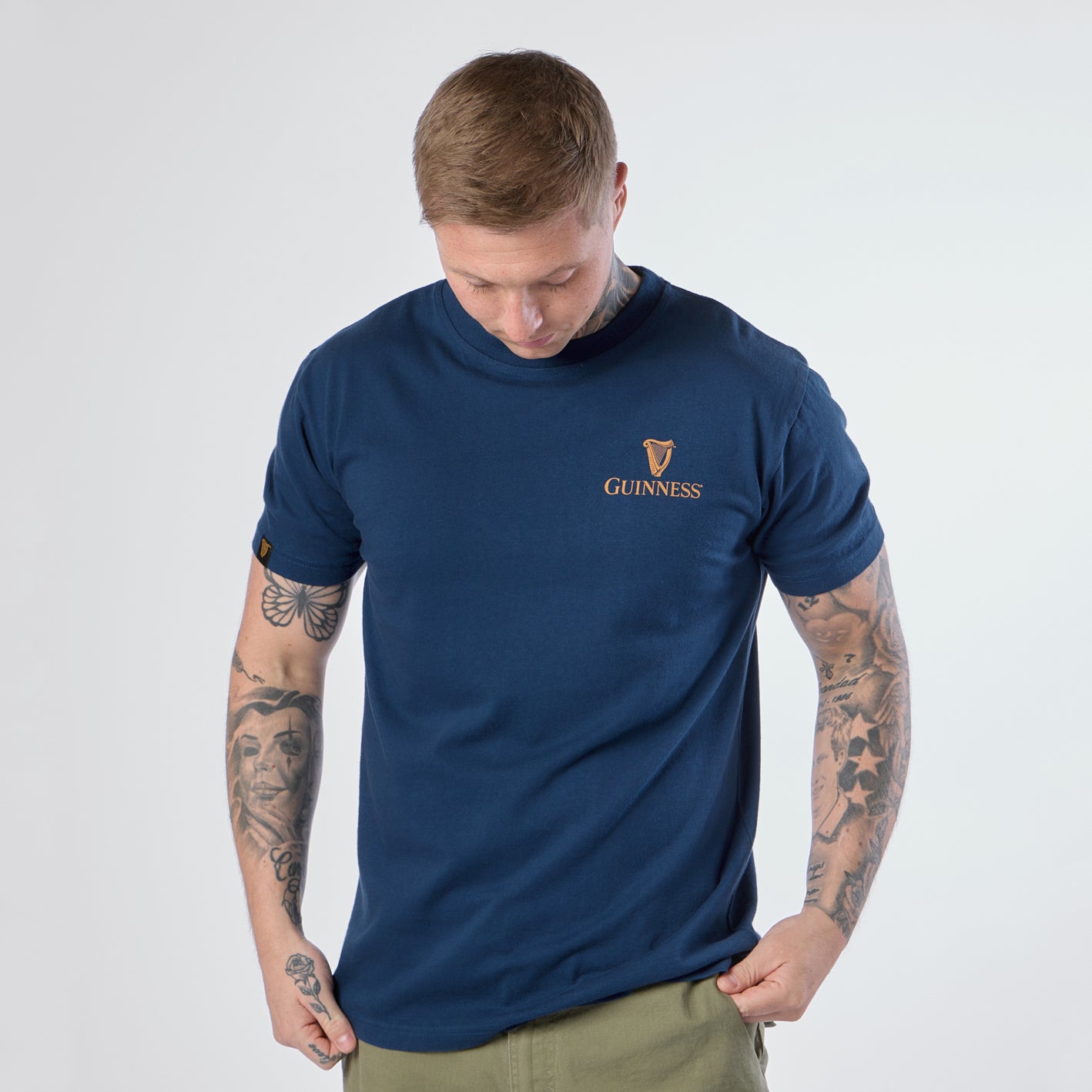 A person with tattoos and short hair looks down while wearing the Guinness Gilroy Seal Tee in blue from Guinness Webstore US and light green pants against a plain background.