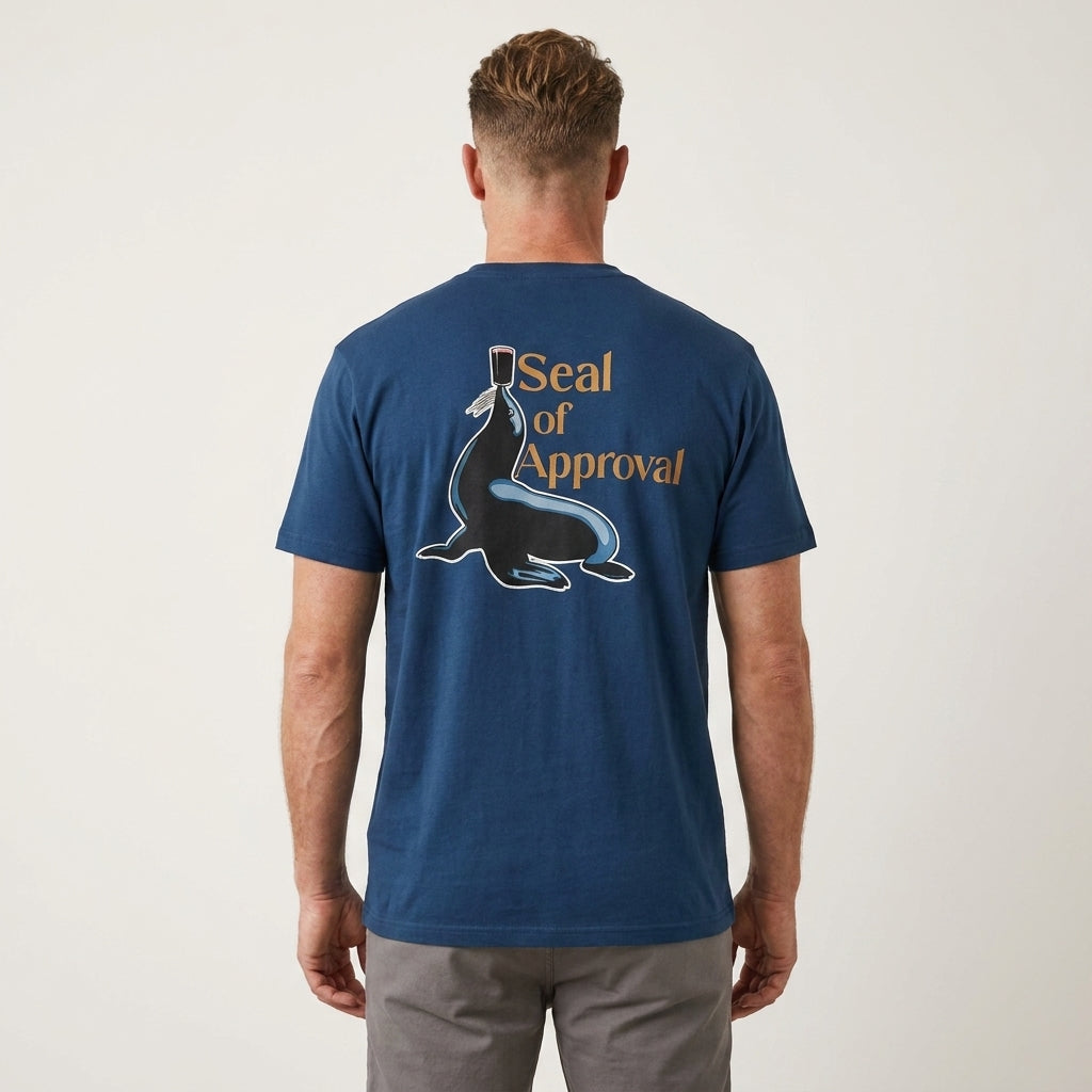 A man in a Guinness Gilroy Seal Tee – Blue from Guinness Webstore US, featuring a seal illustration with a pint of Guinness balanced on his nose and "Seal of Approval" text on the back, stands against a plain background.