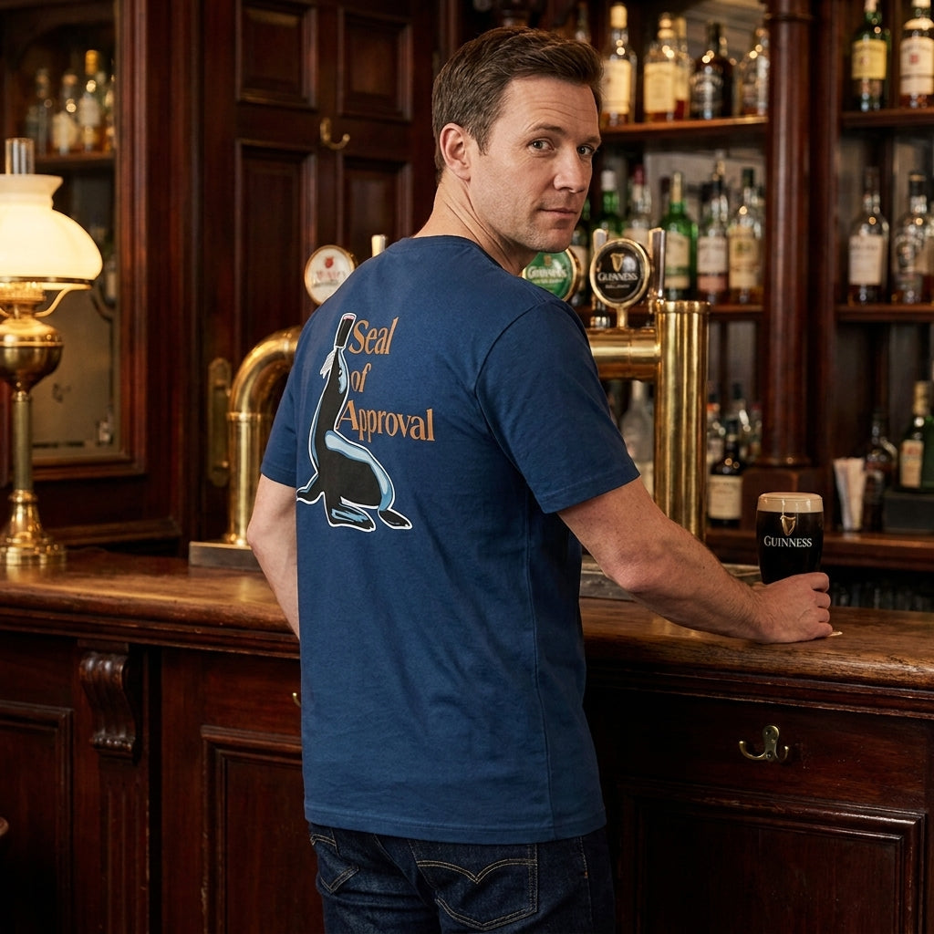 A man at a bar holds a pint of Guinness, wearing the Guinness Gilroy Seal Tee – Blue from Guinness Webstore US, featuring a "Seal of Approval" graphic inspired by the classic Gilroy collection.