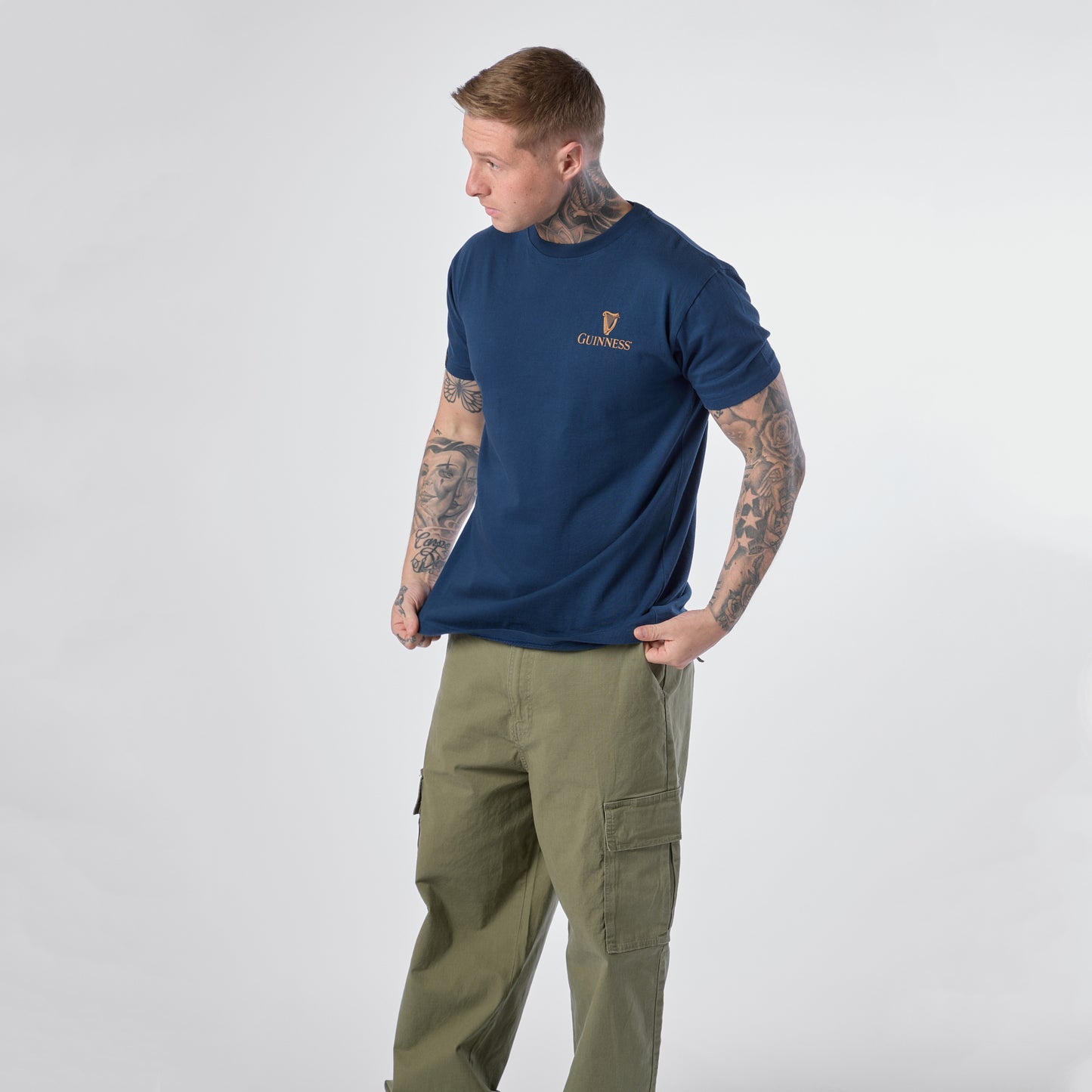 A tattooed individual wearing the Guinness Gilroy Seal Tee – Blue from the iconic Gilroy collection by Guinness Webstore US, paired with olive cargo pants, stands against a plain light background and looks to their left.