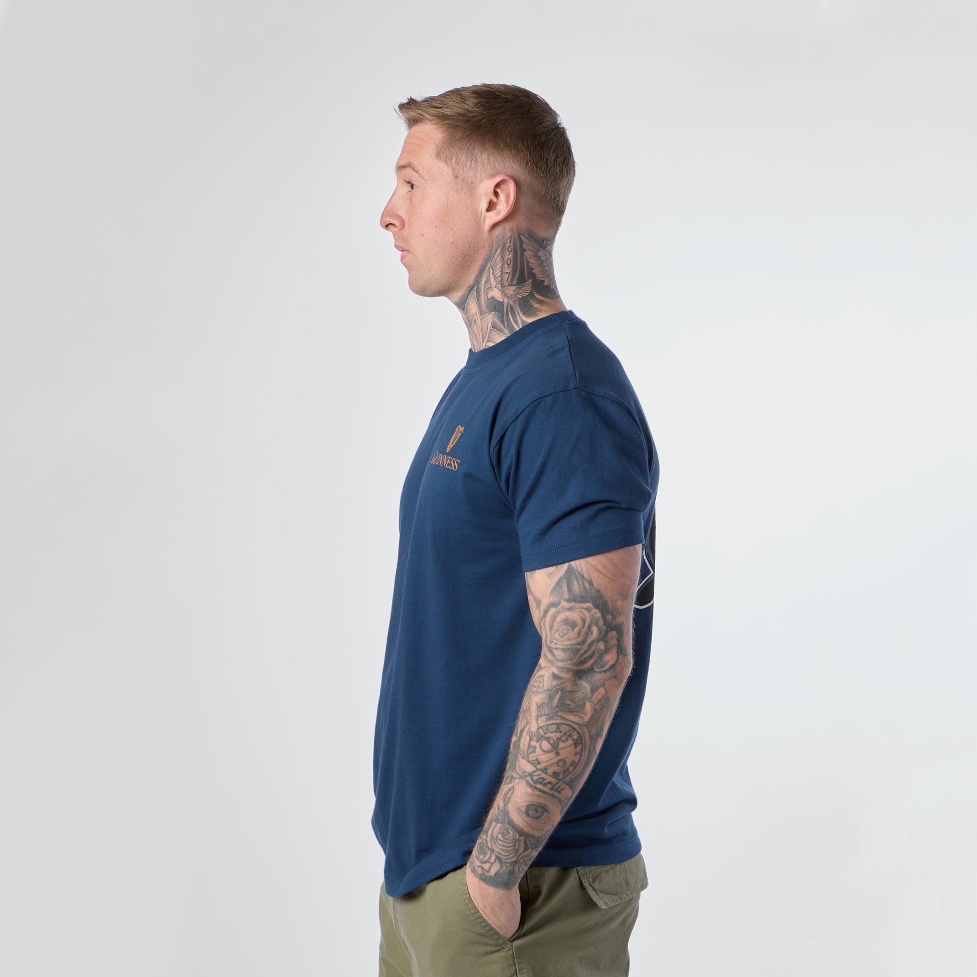 A man with tattoos on his neck and arms stands in profile, wearing a blue Guinness Gilroy Seal Tee from Guinness Webstore US and olive green pants against a plain white background.
