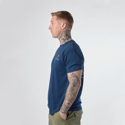 A man with tattoos on his neck and arms stands in profile, wearing a blue Guinness Gilroy Seal Tee from Guinness Webstore US and olive green pants against a plain white background.